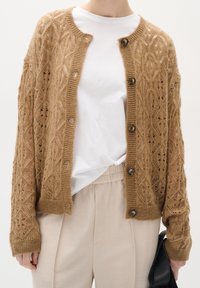 Gebreid vest in zandkleur met een geometrisch ajourpatroon, voorzien van knopen aan de voorkant en geribbelde manchetten, gedragen over een wit overhemd en beige broek.