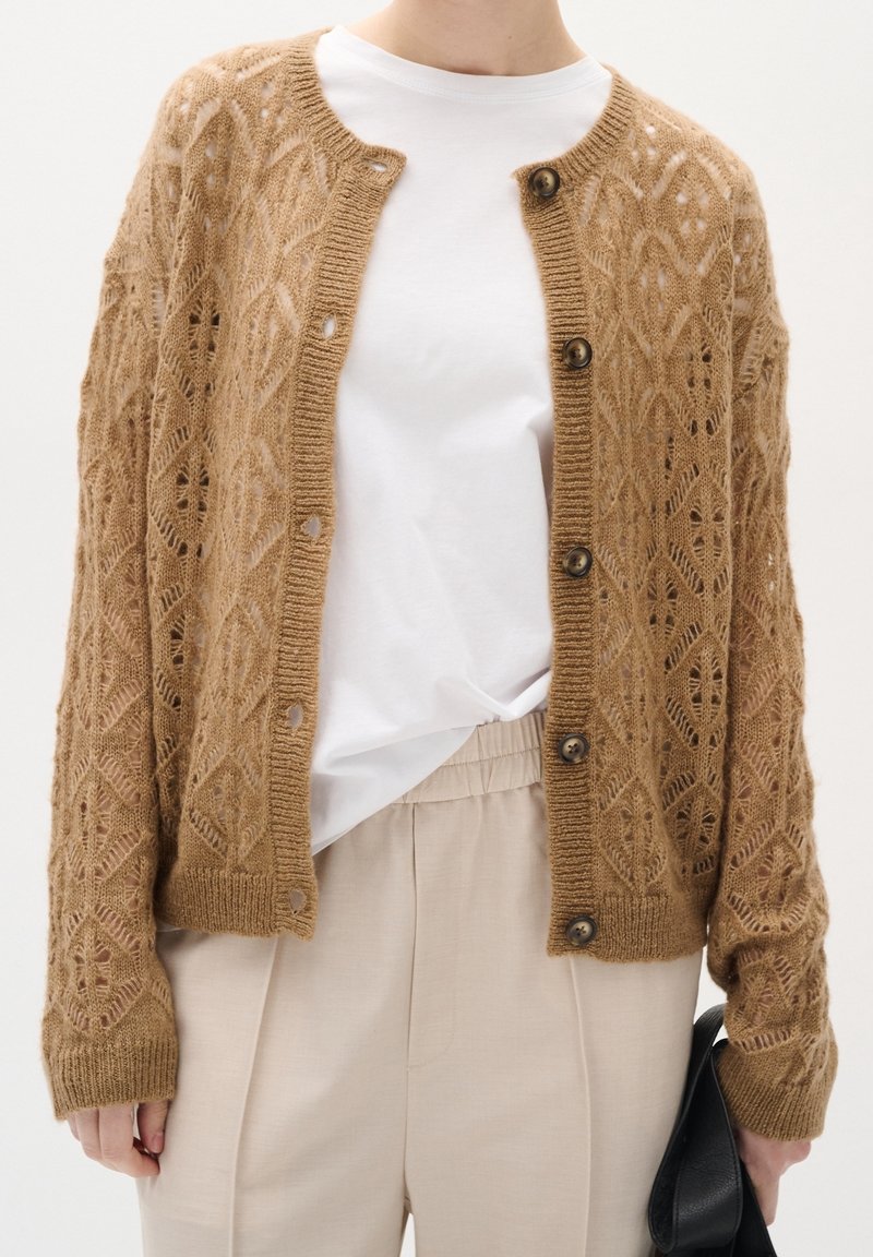 Gebreid vest in zandkleur met een geometrisch ajourpatroon, voorzien van knopen aan de voorkant en geribbelde manchetten, gedragen over een wit overhemd en beige broek.