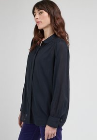 Blusa blu navy a maniche lunghe realizzata in un tessuto leggero; presenta un colletto classico, chiusura a bottoni e spalle leggermente arricciate.