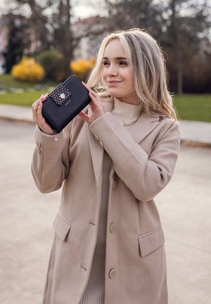 Schwarze Geldbörse mit Reißverschluss und Klappe mit Punktemuster, ausgestattet mit einem Druckknopf. Sie hat eine strukturierte Oberfläche und wird von einer Person getragen, die einen beigen Mantel trägt.