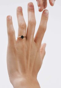 Anillo de oro con una piedra negra cuadrada, con una banda delgada y un diseño geométrico, llevado en una mano de tono de piel claro.