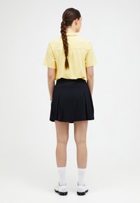 Polo shirt giallo a maniche corte abbinato a una gonna plissettata nera. L'outfit è completato da calze bianche e scarpe nere.