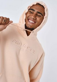 Licht perzik kleurige hoodie met een kangoeroezak, trekkoordkap en een reliëf "PROJECT X" logo op de borst. Zachte stof met een gladde textuur.