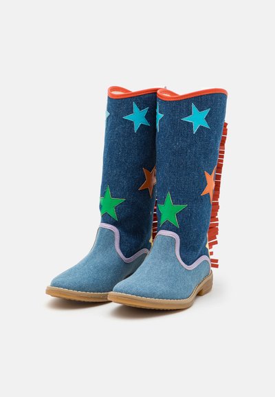 Høye denimbukser med flerfargede stjerneapplikasjoner, oransje frynser på baksiden, lilla kant nær tærne, og lysebrune såler.