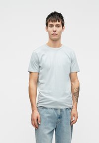 Világoskék pamut póló, kerek nyakkal, rövid ujjal és slim fit fazonnal. Elmosódott kék farmernadrággal párosítva. Egyszerű dizájn, látható logók nélkül.