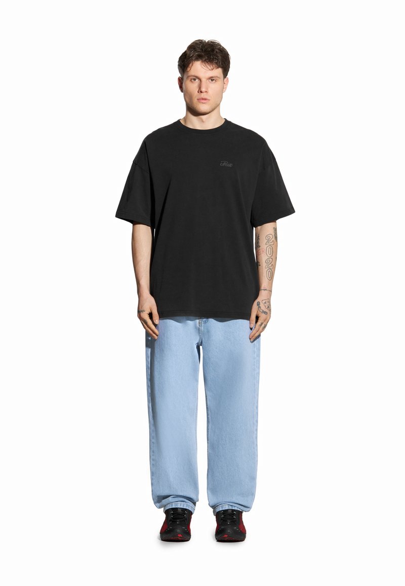 Czarna koszulka z krótkim rękawem z subtelnym logo, zestawiona z jasnoniebieskimi luźnymi dżinsami i czarnymi trampkami z czerwonymi akcentami.