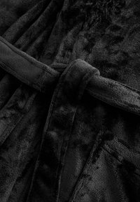 Juodas veliūrinis chalatas su minkšta tekstūra, su pririštu diržu ir dviem priekinėmis kišenėmis. Audinys turi glotnią, aksominę išvaizdą.