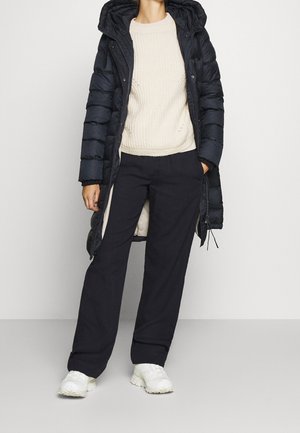 Personne portant un manteau matelassé à capuche noir sur un pull crème, un pantalon large noir et des baskets blanches, debout devant un fond uni.