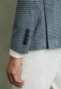 Sacou cu tipar în carouri albastre, din țesătură texturată, cu manșetă cu patru nasturi și mânecă de cămașă albă care se zărește dedesubt.