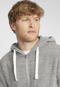 Sivi zip-up hoodie s mekim teksturom, sa vezicama i kapuljačom. Vidljivi detalji šavova; nosi se preko bijelog T-shirta.