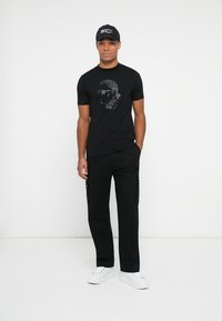 Camiseta negra con un gráfico de cara geométrica, combinada con pantalones cargo negros y zapatillas blancas. Presenta una gorra negra con acentos de texto.