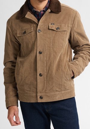 Mężczyzna w brązowej kurtce sztruksowej z ciemnobrązowym kołnierzem z baranka, zapinanej na guziki, z klapami na kieszeniach piersiowych i ciemnoniebieskich dżinsach.