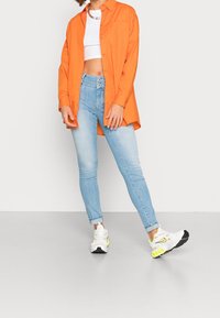 Camisa de botones naranja sobre un top corto blanco, combinada con unos jeans ajustados de color azul claro y zapatillas blancas con acentos amarillos.