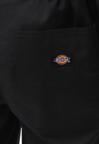 Černé pracovní kalhoty vyrobené z odolné látky, vybavené náprsní kapsou s barevným logem "Dickies" na zadní straně.