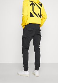 Zwarte cargobroek met zij- en achterzakken, gedragen met een felgele hoodie met zwarte grafische ontwerpen, gecombineerd met witte sneakers.