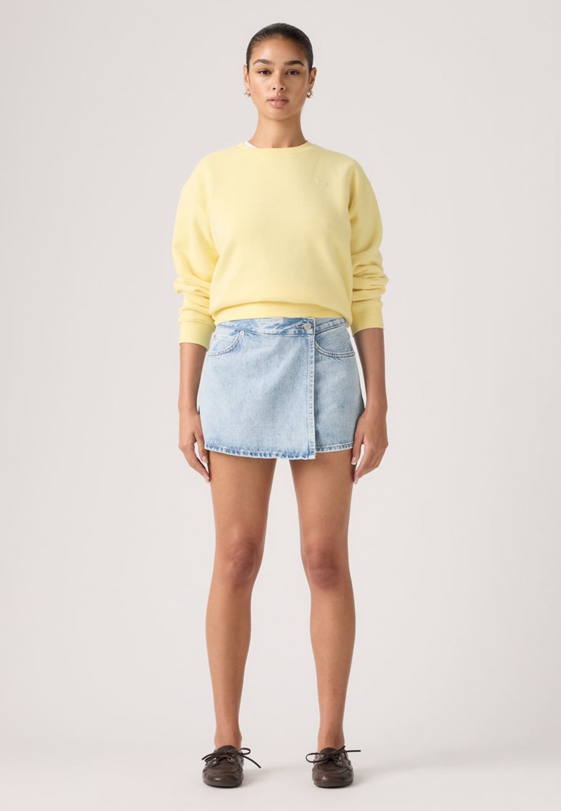 Νεαρή γυναίκα όρθια, φοράει ένας ανοιχτό κίτρινο φούτερ με μακριά μανίκια, μίνι φούστα από ανοιχτό μπλε τζιν και σκούρα καφέ παπούτσια με κορδόνια.