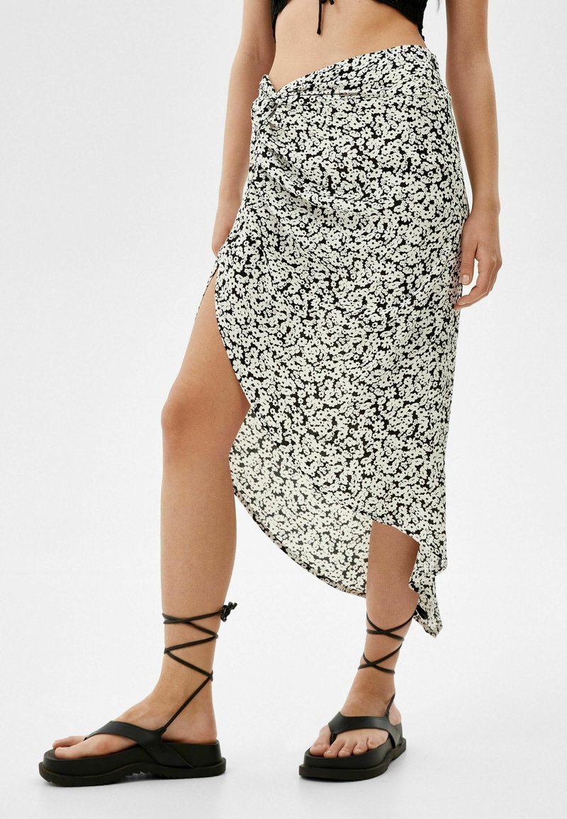 Hame panjang berbunga hitam dan putih dengan belahan tinggi, menampilkan detail depan yang terikat. Dipadukan dengan sandal strappy hitam.