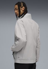 Chaqueta cortavientos gris claro con un cuello texturizado, dos bolsillos laterales y un ajuste relajado. Presenta herrajes de marca cerca del dobladillo.