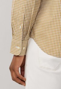 Camisa de cuadros en beige y blanco con puños abotonados. Presenta un corte relajado y un dobladillo redondeado. Se lleva con pantalones de color claro.