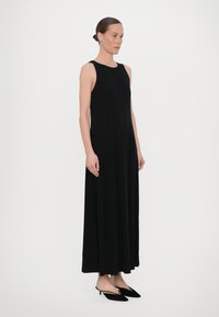 Czarna maxi sukienka bez rękawów, wykonana z miękkiego materiału, charakteryzująca się luźnym krojem i gładką fakturą, zestawiona z eleganckimi czarnymi szpilkami.