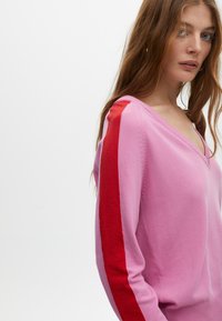 V-Ausschnitt-Pullover in Hellrosa mit roten und dunkelpinken Streifen an den Ärmeln. Weiche Textur, figurbetontes Design und geripptes Saumdetail.