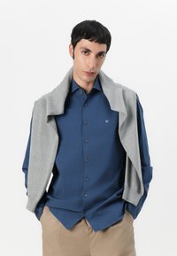 Chemise bleu à boutons avec un pull gris drapé sur les épaules. La chemise a un léger éclat, et le pull présente des poignets côtelés.
