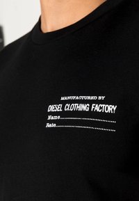 Svart bomullströja med en vit tryckt etikett med texten "TILLVERKAD AV DIESEL CLOTHING FACTORY" och rader för "Namn" och "Bedömning."