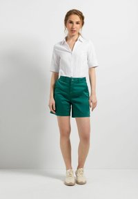 Weißes kurzärmeliges Hemd mit Knopfleiste, kombiniert mit grünen Shorts. Die Shorts haben eine flache Vorderseite mit Knopfverschluss und seitlichen Taschen.