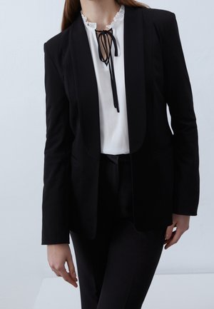 Femme portant un blazer noir taillé sur mesure et un pantalon, avec un chemisier blanc à col volanté et un nœud noir au niveau du col.