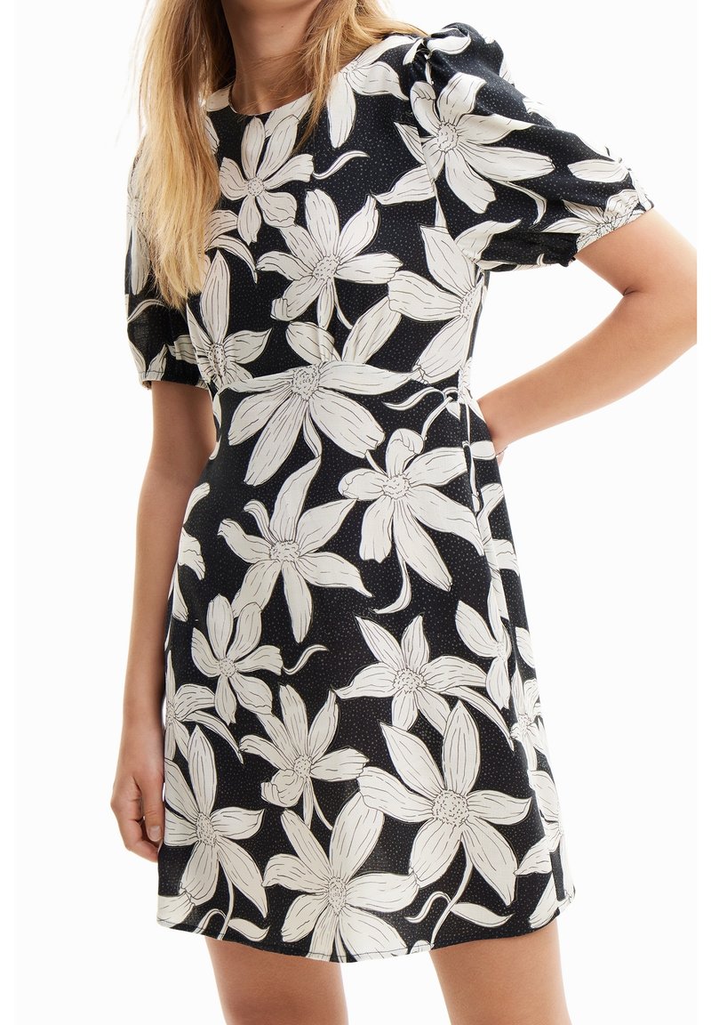 Femme portant une robe noire à grand imprimé floral blanc avec des manches courtes bouffantes, posant avec une main sur la hanche.