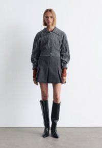 Grauer Kabelstrick-Cardigan mit kragen, kombiniert mit einem dunkelgrauen, gefältelten Minirock. Abgerundet mit knielangen, schwarzen, krokodilgeprägten Stiefeln.