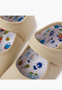 Beige Canvas-Schuhe mit flachem Design, ausgestattet mit einem Klettverschluss und einem bunten, gemusterten Innenfutter mit cartoonhaften Robotern und Fahrzeugen.