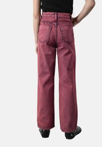 Szerokie dżinsy w wyblakłym czerwonawym kolorze, z wysokim stanem, klasyczną fakturą dżinsu oraz dwiema tylnymi kieszeniami. Czarne buty z widocznymi dziurkami.