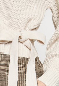 Béžový pletený svetr s texturovaným povrchem a páskem vázaným na mašli. Nositelný přes vzorované hnědé kárované kalhoty.