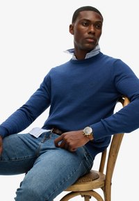 Blå strikket sweater med rund hals, lagd ovenpå en lyseblå og hvid stribet skjorte. Båret med denim jeans og et brunt armbåndsur.