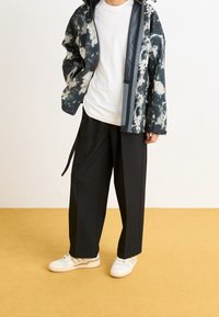 Czarny i biały kurtka tie-dye, biała koszulka, szerokie czarne spodnie i białe sneakersy z beżowymi akcentami, na tle żółtej podłogi.