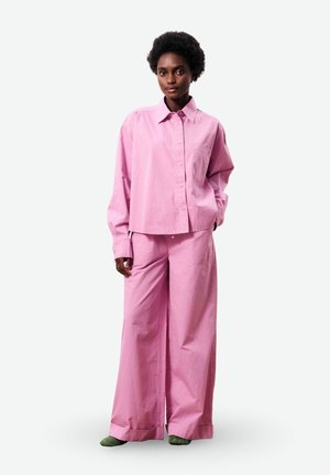 Vrouw met kort krullend haar, gekleed in een losse roze blouse met knopen en wijde roze broek, met groene schoenen.