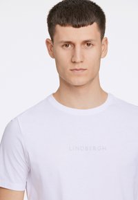 Valkoinen puuvillapaita, jossa on pyöreä kaula-aukko ja hillitty "LINDBERGH" logo kaiverrettuna harmaana rinnassa. Sujuva tekstuuri ja lyhyet hihat.