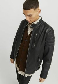 Svart läderjacka med dragkedja framtill, vadderade axlar och sidofickor. Bärs över en brun cardigan och en randig skjorta.