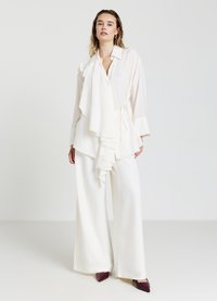 Blusa bianca con frontale arricciato e vita con lacci, abbinata a pantaloni bianchi a gamba ampia. Il look è completato da tacchi a punta bordeaux.