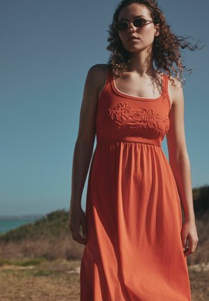 Frau mit lockigem Haar, die eine Sonnenbrille und ein ärmelloses orangenes Kleid mit Blumengstickerei trägt, steht draußen vor klarem Himmel.