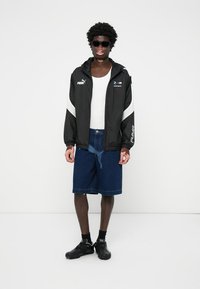 Jachetă windbreaker neagră cu accente albe, combinată cu pantaloni scurți din denim până la genunchi și adidași negri. Modelul poartă ochelari de soare.