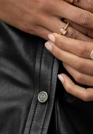 Svart läderjacka med textilstruktur, prydd med en glansig knapplapp. Två ringar pryder fingrar med ljusa naglar.