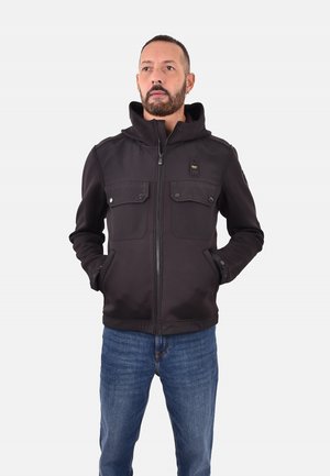 Giacca nera con zip e cappuccio, dotata di due tasche frontali, dettagli con bottoni e una texture morbida. Indossata con jeans blu.