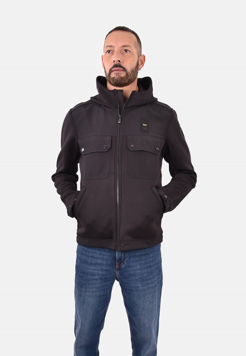 Zwarte zip-up jack met capuchon, voorzien van twee voorzakken, knopenaccenten en een gladde textuur. Draagt met blauwe spijkerbroek.