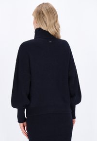 Maglione blu navy con colletto alto, texture a coste e maniche oversize. Presenta un dettaglio del logo minimale sul retro.
