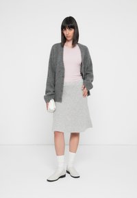 Šedý pletený cardigan, svetloružový top, šedá nadýchaná sukňa, biele ponožky a biele topánky, drží malú bielu tašku. Jednoduchý dizajn, jemné textúry.