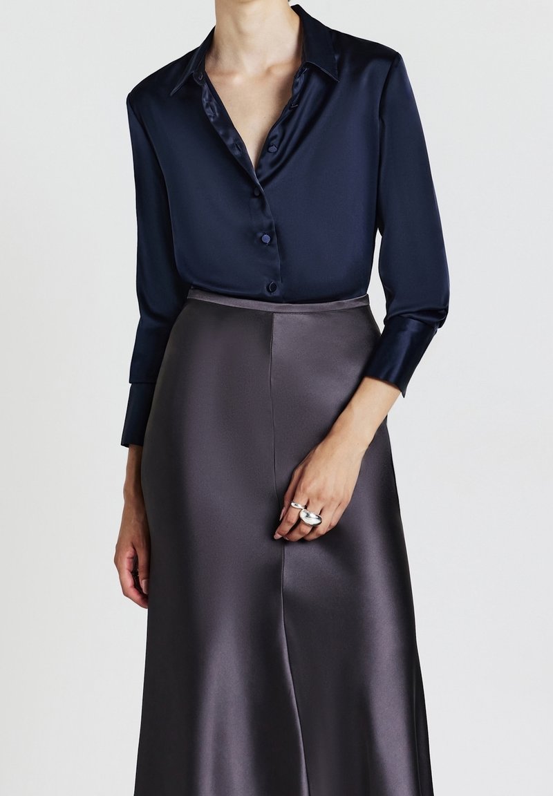 Personne portant une chemise en satin bleu marine foncé boutonnée avec un col et une jupe en satin gris foncé taille haute, montrant une main avec des bagues en argent.