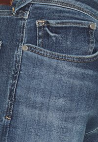 Primer plano de un bolsillo trasero de unos jeans de mezclilla azul con costuras visibles, remaches y textura desgastada.