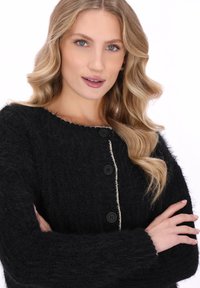 Zwart gebreid vest met een fluweelachtige textuur, voorzien van een ronde halslijn, grote zwarte knopen en een contrasterende lichte bies langs de voorkant.